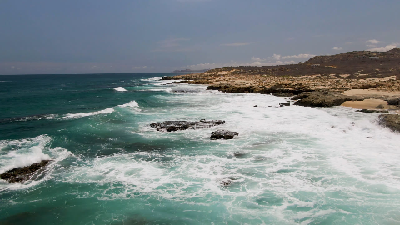 Wide view of Laguna Beach in Baja California Sur with white surf spreading over dark rocks, tan sand, dry hills, and no visible people.