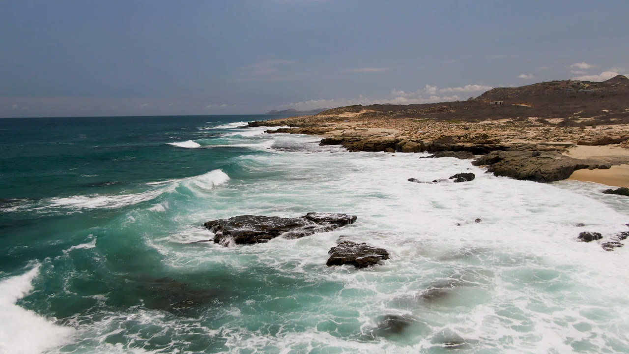 Laguna Beach shoreline with a large white wave line crossing the foreground, exposed rock shelves in the middle, distant hills, and no visible people.