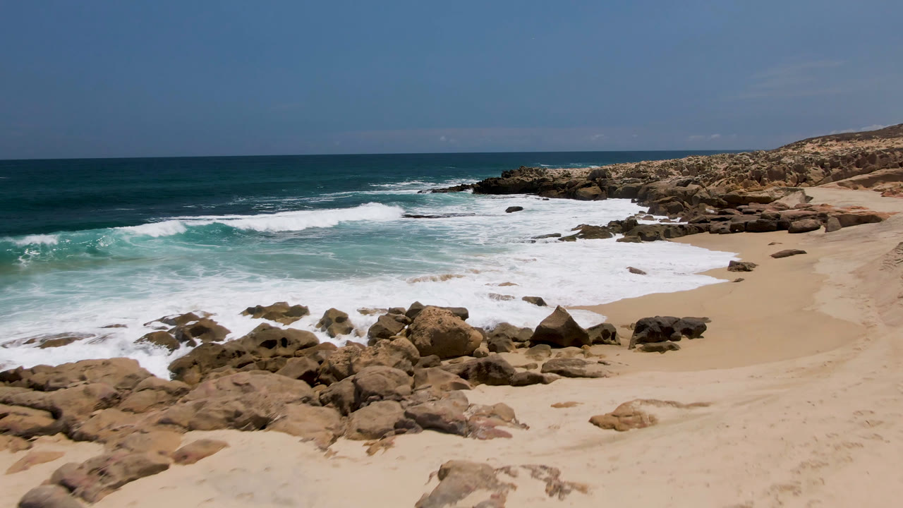 Laguna Beach shoreline in Baja California Sur with more white foam spreading across the sand, dark rocks in the foreground, a rocky headland, and no visible people.