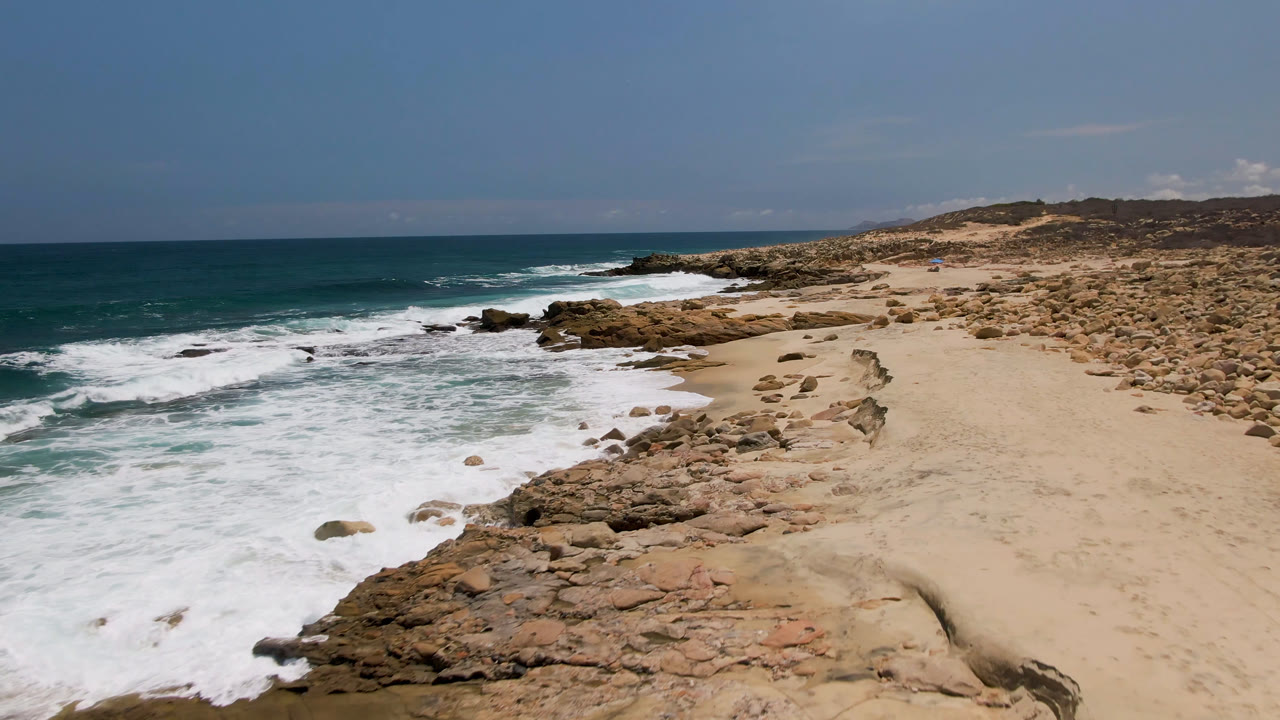 Opening frame over Laguna Beach, Baja California Sur, showing white surf rolling onto a rocky sand beach with no visible people.