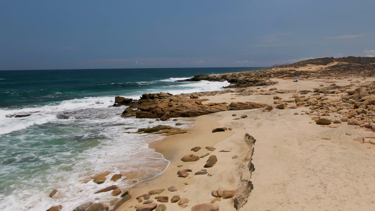 White waves curve onto sand near scattered rocks and dry coastal hills.