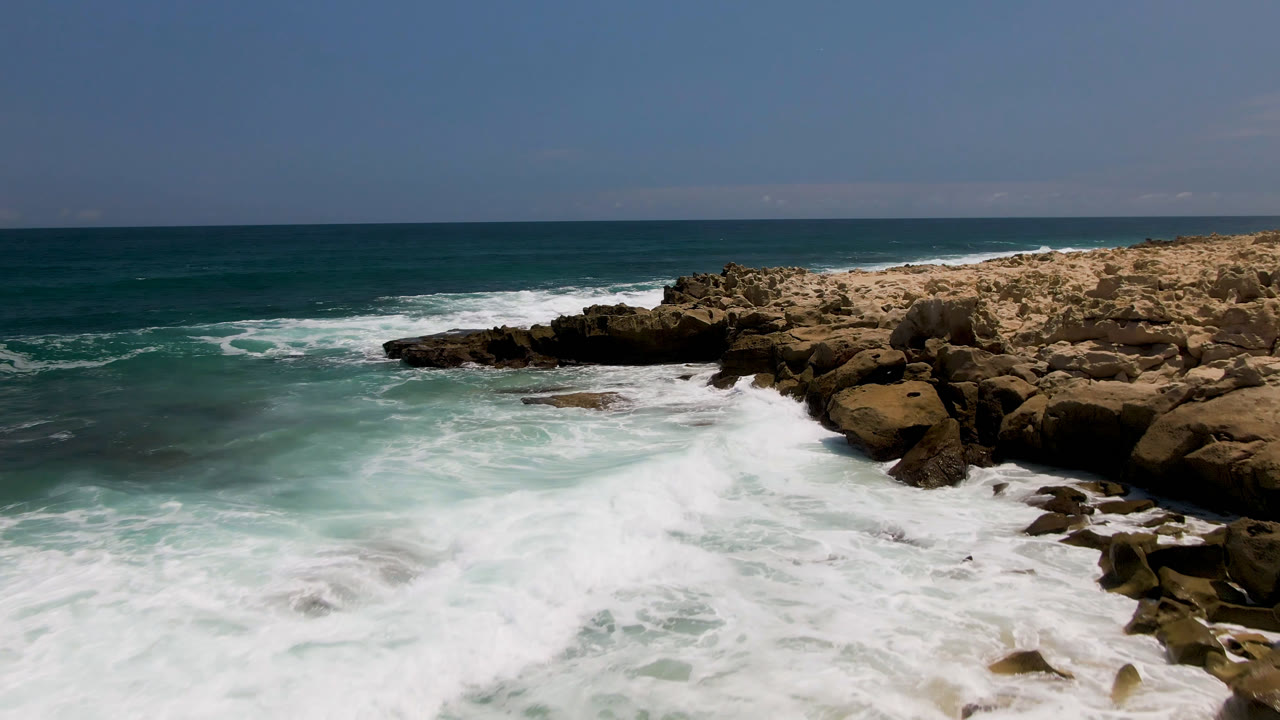 At Laguna Beach in Baja California Sur, white surf breaks around beige rock shelves and dark outcrops under a clear blue horizon with no visible people.