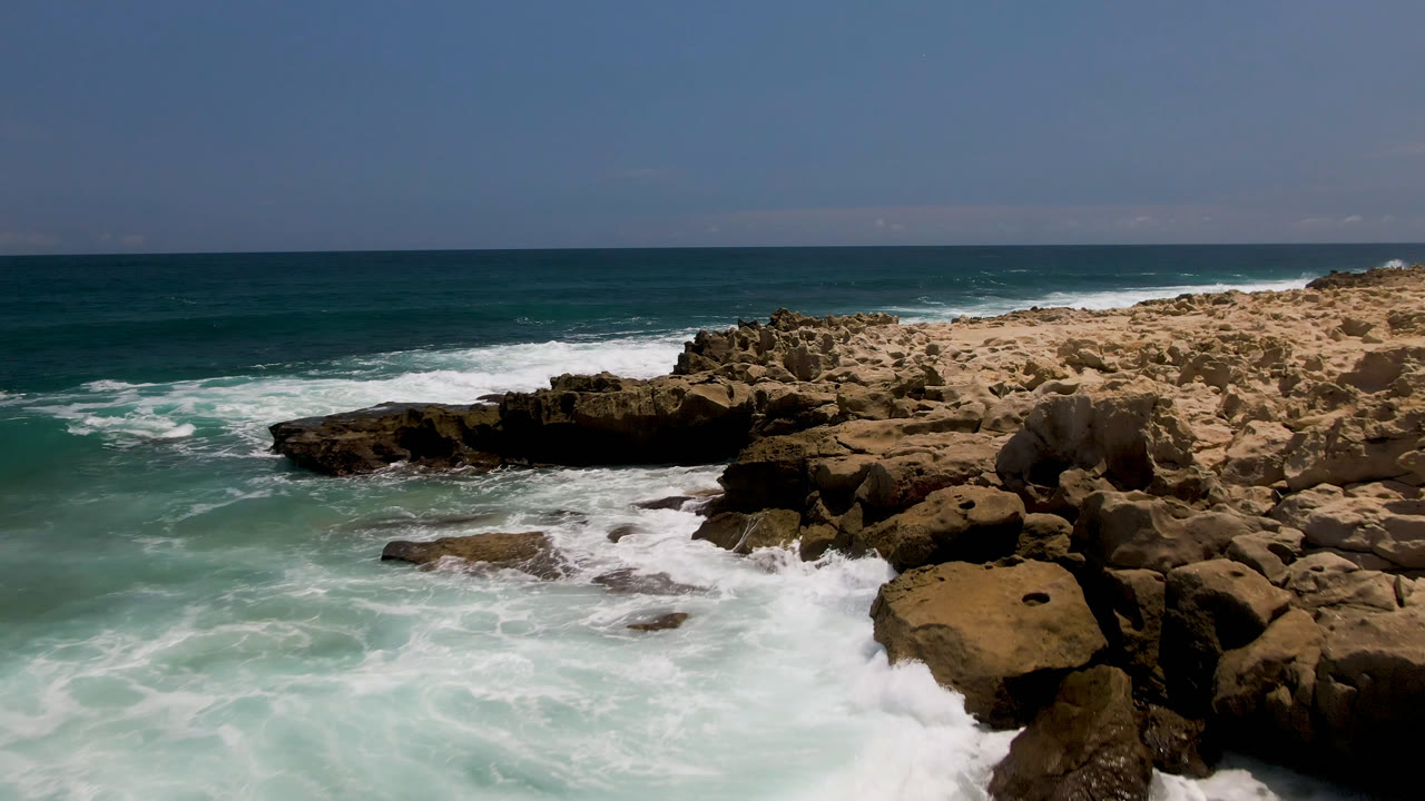 At Laguna Beach in Baja California Sur, a low rock point extends into teal water as foamy waves wrap around both sides with no visible people.