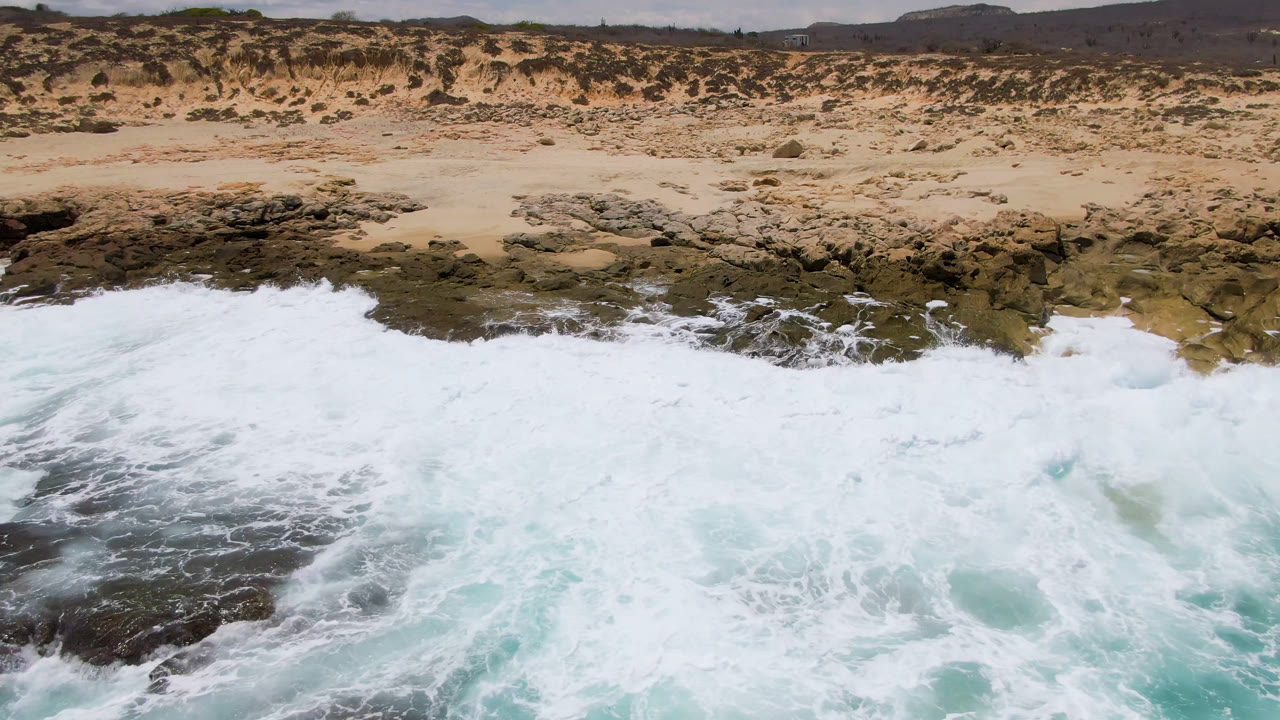 Breaking surf reaches a rocky beach below sand and scrubland.