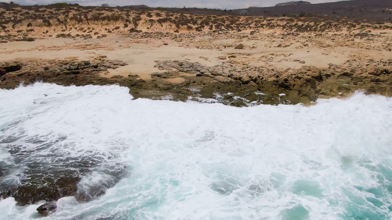A broad sheet of white seafoam pushes farther across the rocky shoreline at Laguna Beach, Baja California Sur, with sand and low vegetation behind it and no visible people.