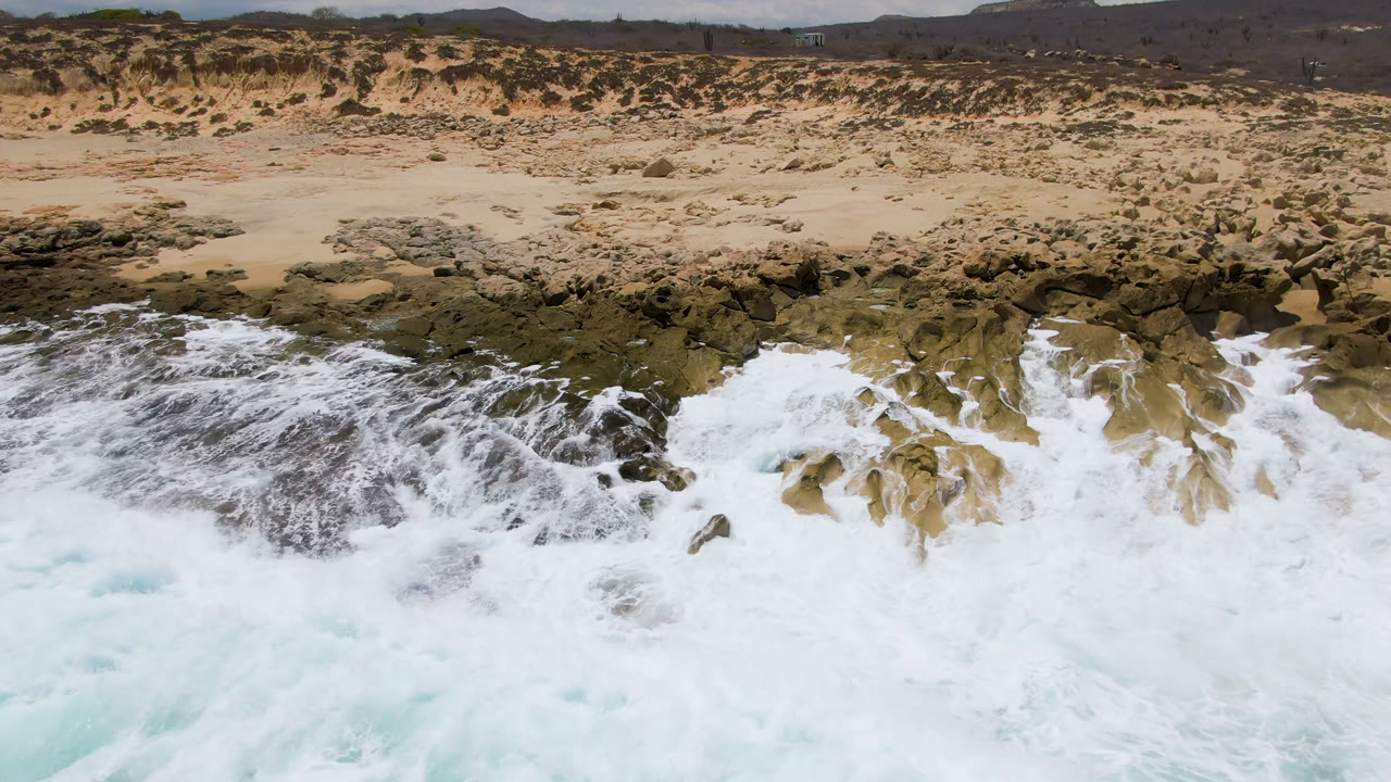Aerial view of white surf breaking across dark rocks at Laguna Beach, Baja California Sur, with tan dunes and low hills beyond and no visible people.