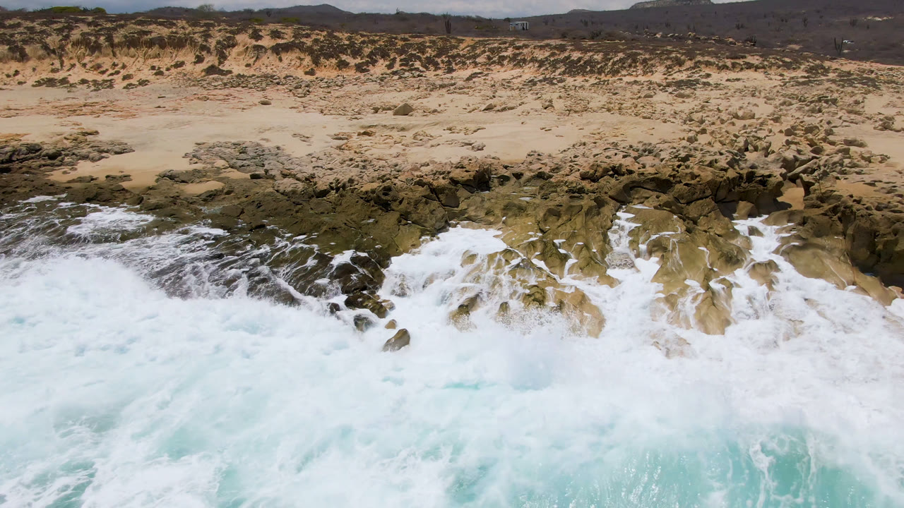 Foam spreads over the rocky reef at Laguna Beach, Baja California Sur, while the sandy beach and scrub-covered dunes remain clear in the background with no visible people.