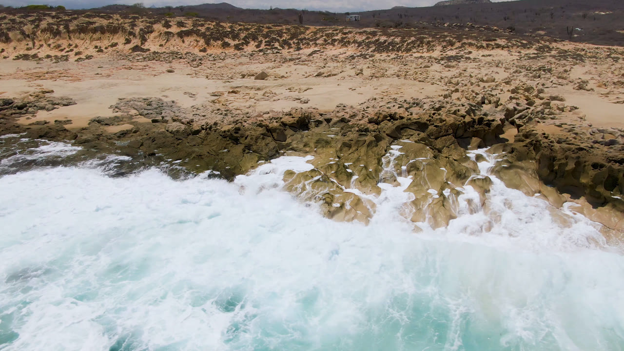 A larger white wave surges into jagged rocks at Laguna Beach, Baja California Sur, with pale blue water below and empty sand and dunes above, no visible people.
