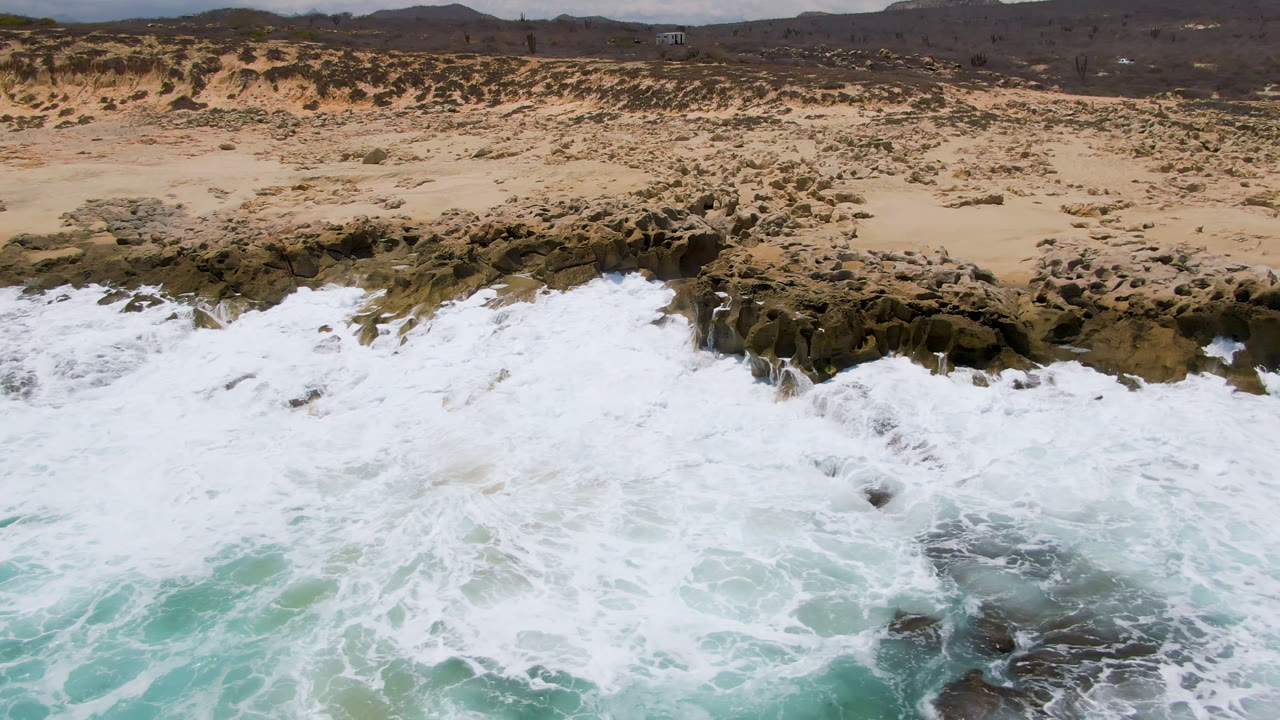 Aerial frame from Laguna Beach, Baja California Sur, Mexico, with white surf spreading across dark shoreline rocks below sandy scrubland and no visible people.