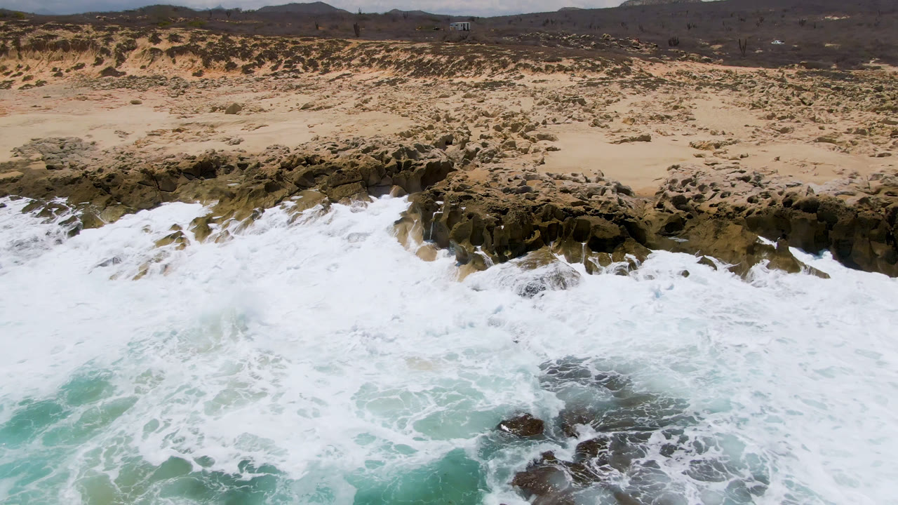 Drone view over Laguna Beach, Baja California Sur, Mexico, as pale turquoise water and white foam cover the lower frame while scrubby sand and hills remain in the background, with no visible people.