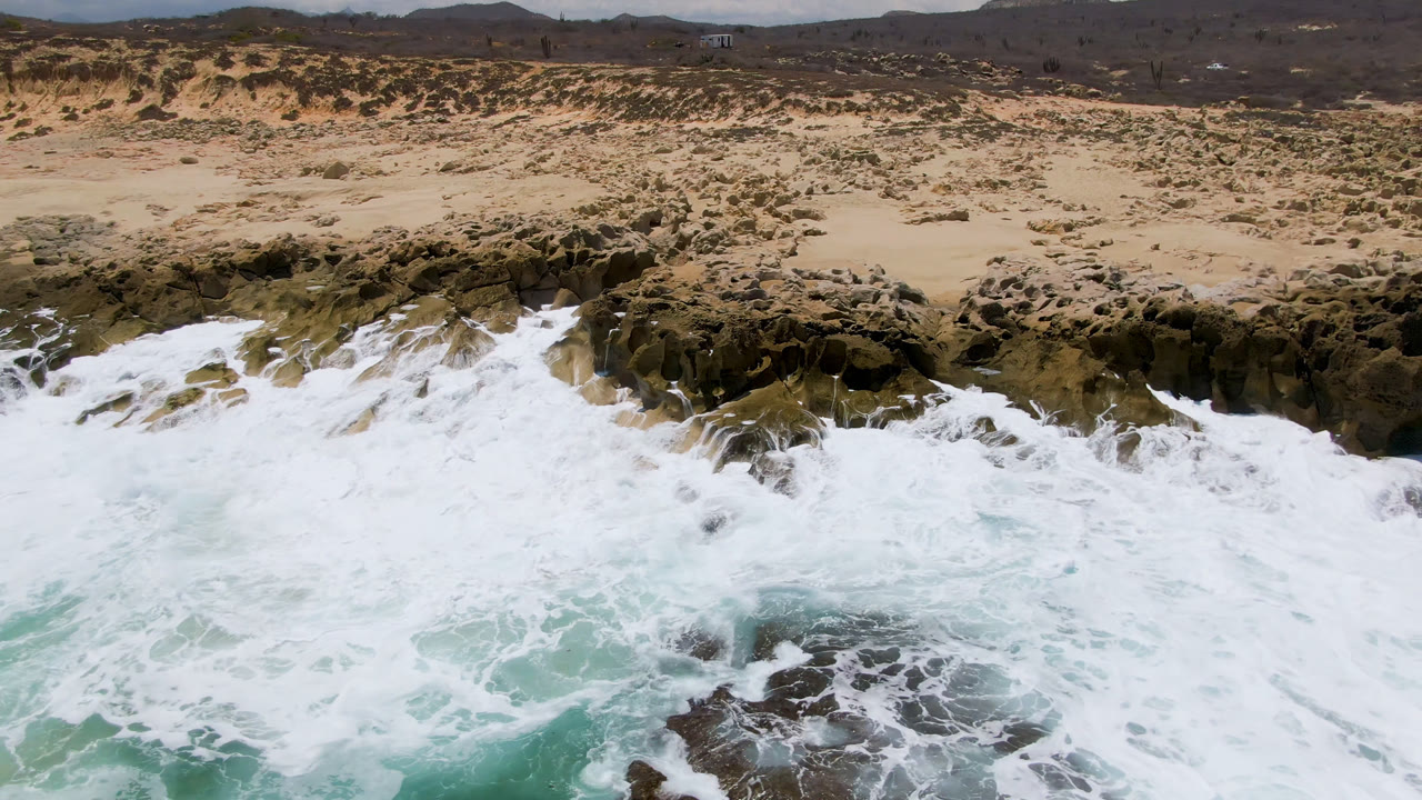 Aerial coastline with surf exploding over dark rocks below dry beach land.