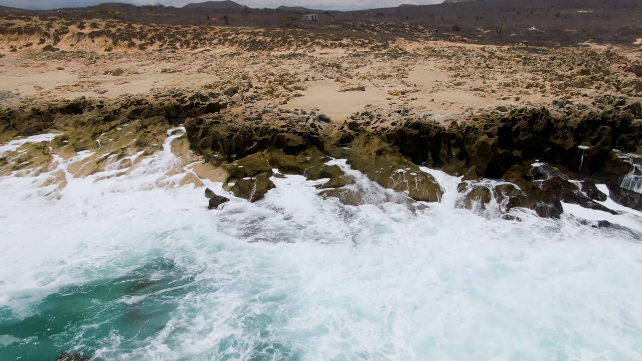 Aerial frame of Laguna Beach, Baja California Sur, with whitewater surging over dark shoreline rocks, pale sand and scrub behind them, distant hills on the horizon, and no visible people.