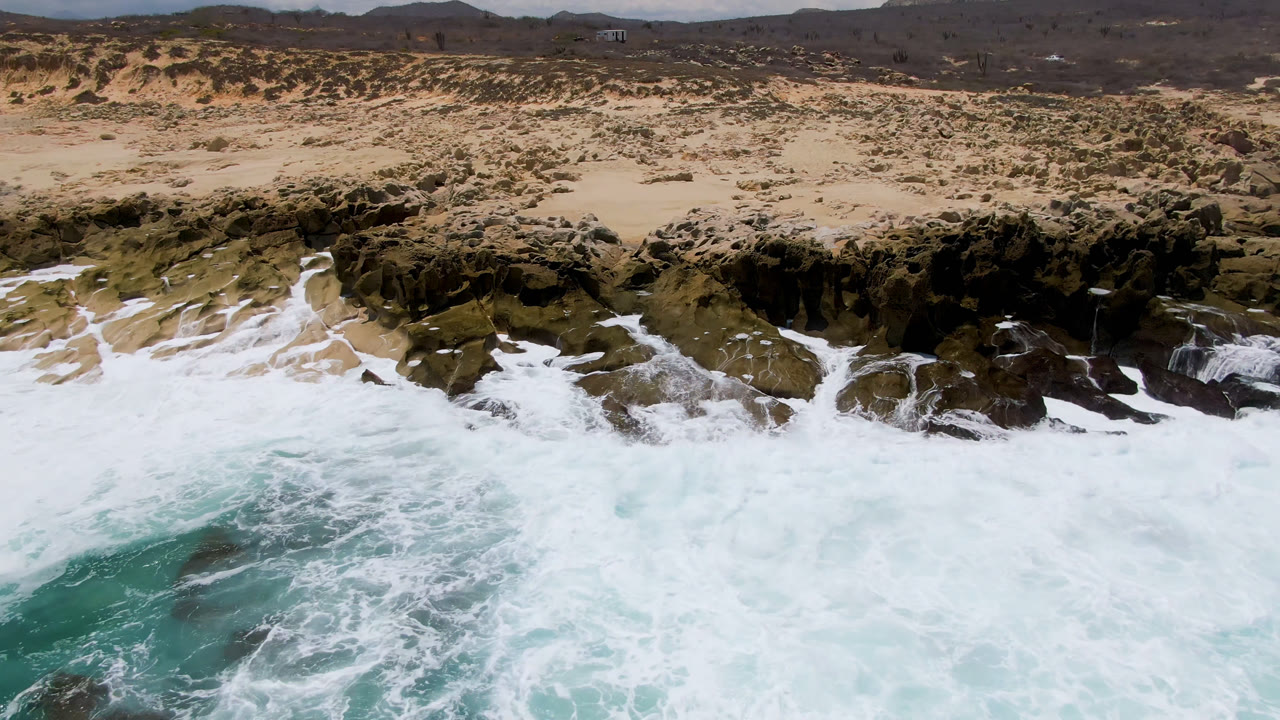 Aerial view at Laguna Beach, Baja California Sur, where turquoise water and white foam spread across dark rocks beneath a sandy, scrub-covered beach with no visible people.