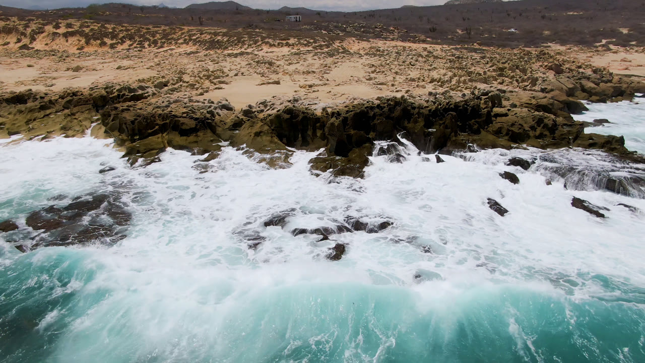 Aerial frame at Laguna Beach, Baja California Sur, Mexico showing turquoise water and white foam breaking over dark rocks, with sandy scrub dunes and no visible people.