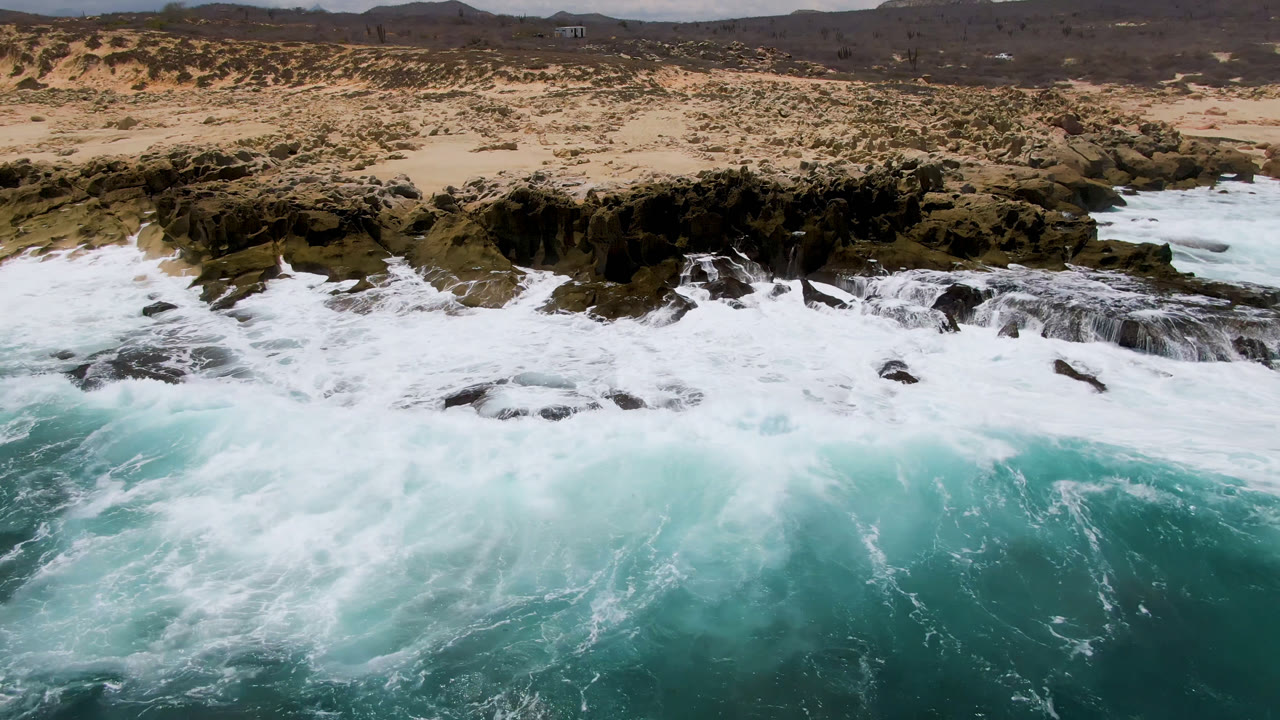 At 0:01, the aerial view shows a wider sheet of white water over the rocks at Laguna Beach, with tan sand, sparse vegetation, and no visible people.