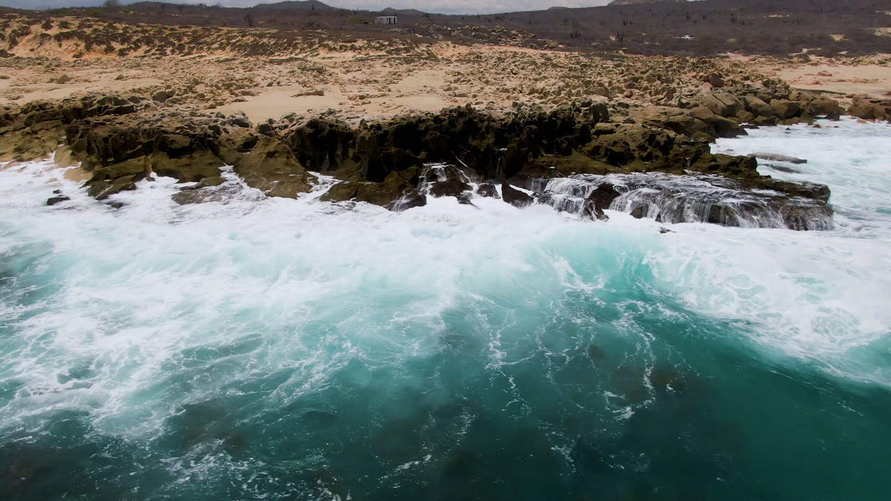 Foamy wave collapses against jagged rocks along a sandy Baja California Sur shore.