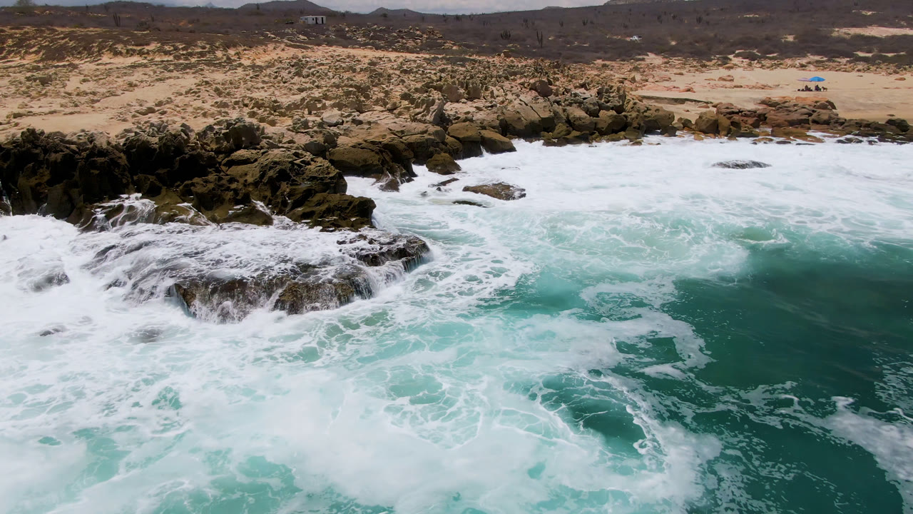 At Laguna Beach in Baja California Sur, white surf wraps around dark rocks with turquoise water in front and sandy scrubland behind.