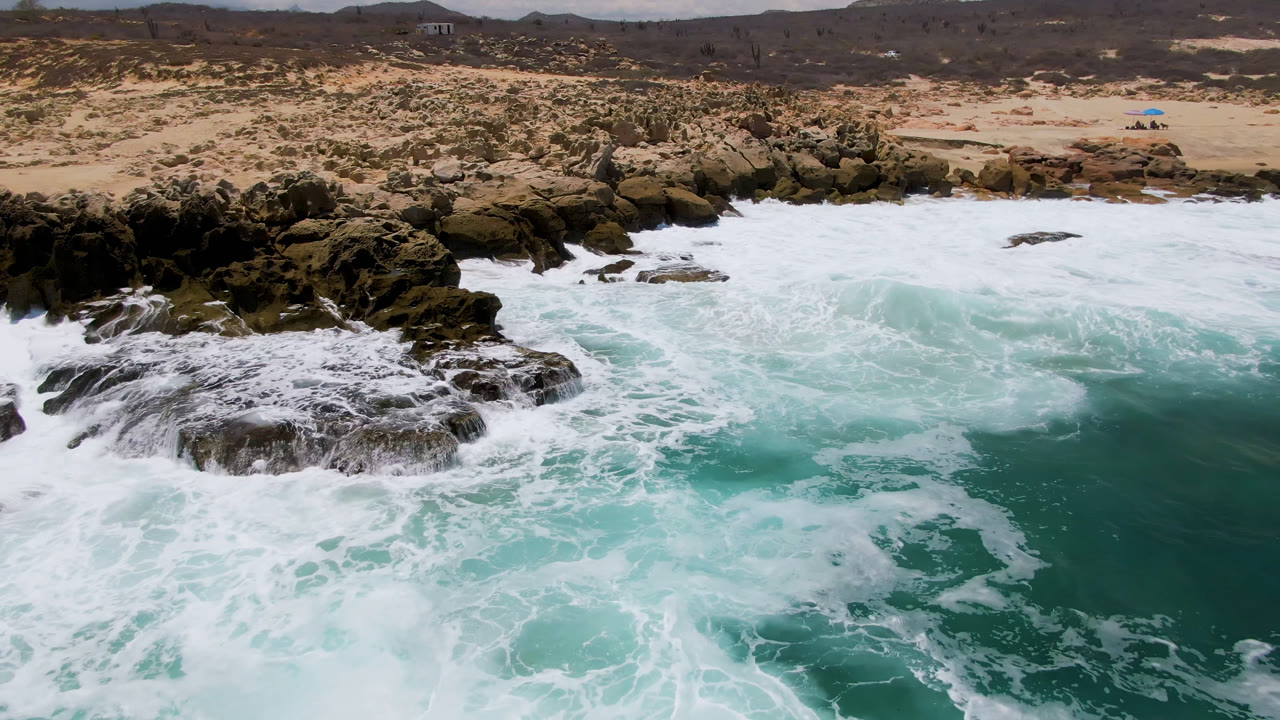 Whitewater spreads over the central rock formation at Laguna Beach, Baja California Sur, with pale sand, scrub, and low hills across the horizon.