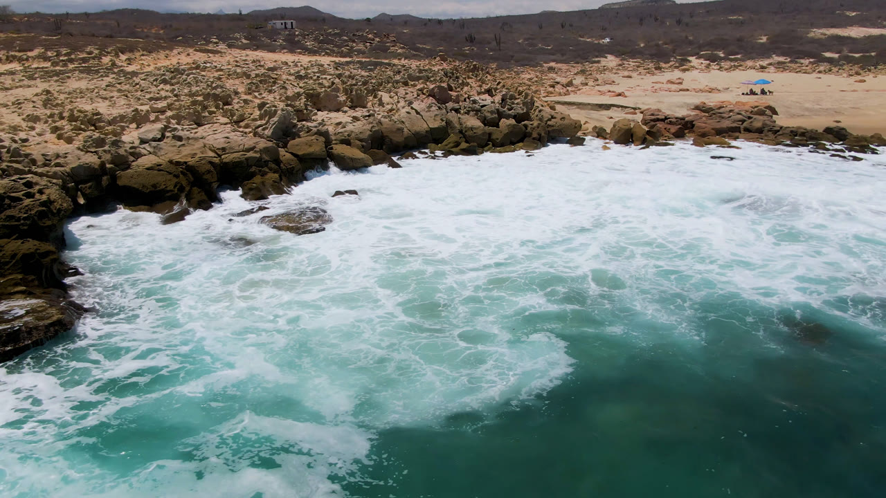 Aerial frame over Laguna Beach, Baja California Sur, with turquoise water, white foam, dark shoreline rocks, sandy beach, desert scrub, distant hills, and no visible people.