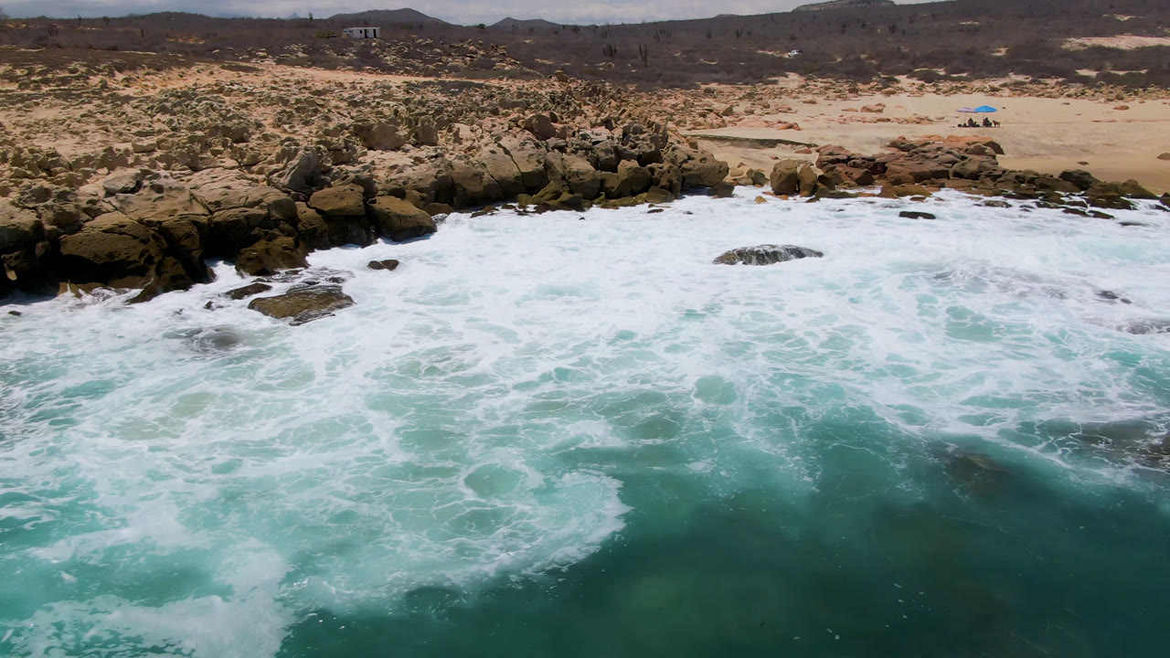 Aerial view at Laguna Beach, Baja California Sur, as white surf thickens across the foreground rocks, with tan sand, sparse scrub, low hills, and no visible people.