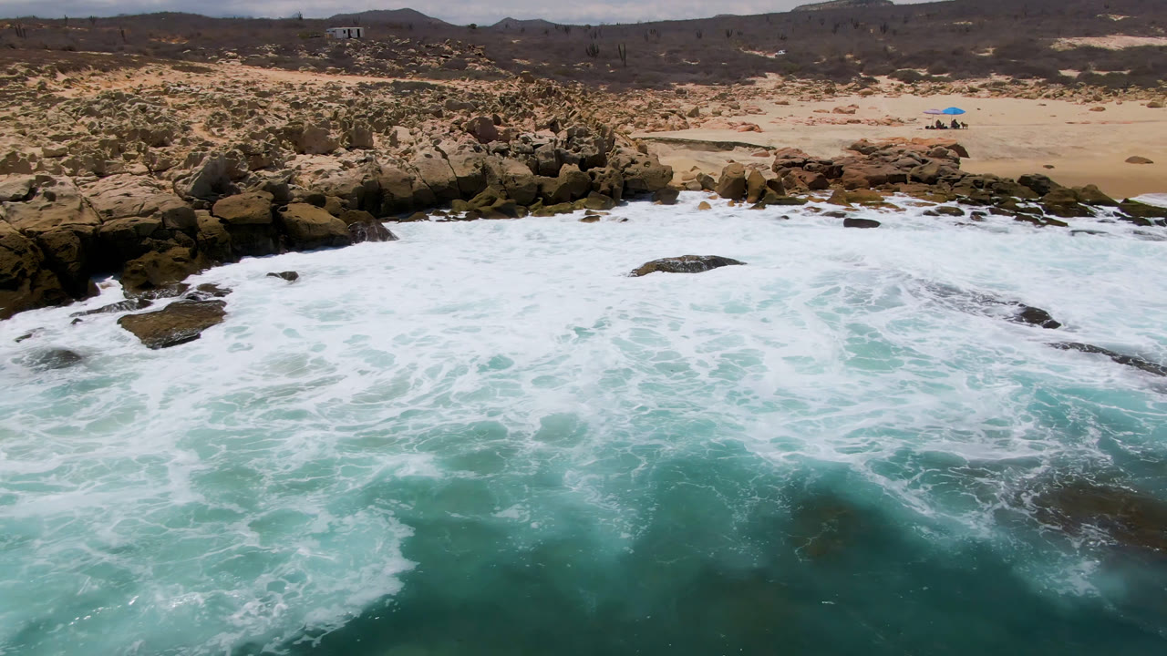 Foamy turquoise water spreads across rocks with a sandy beach in the background.