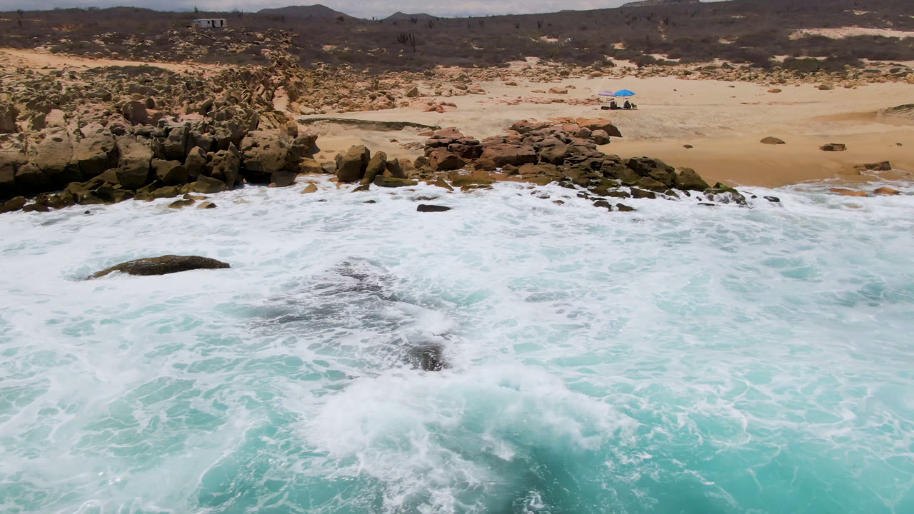 A broad wash of white surf spreads around exposed rocks at Laguna Beach, Baja California Sur, with pale sand, scrub, and a small blue beach object in the background.