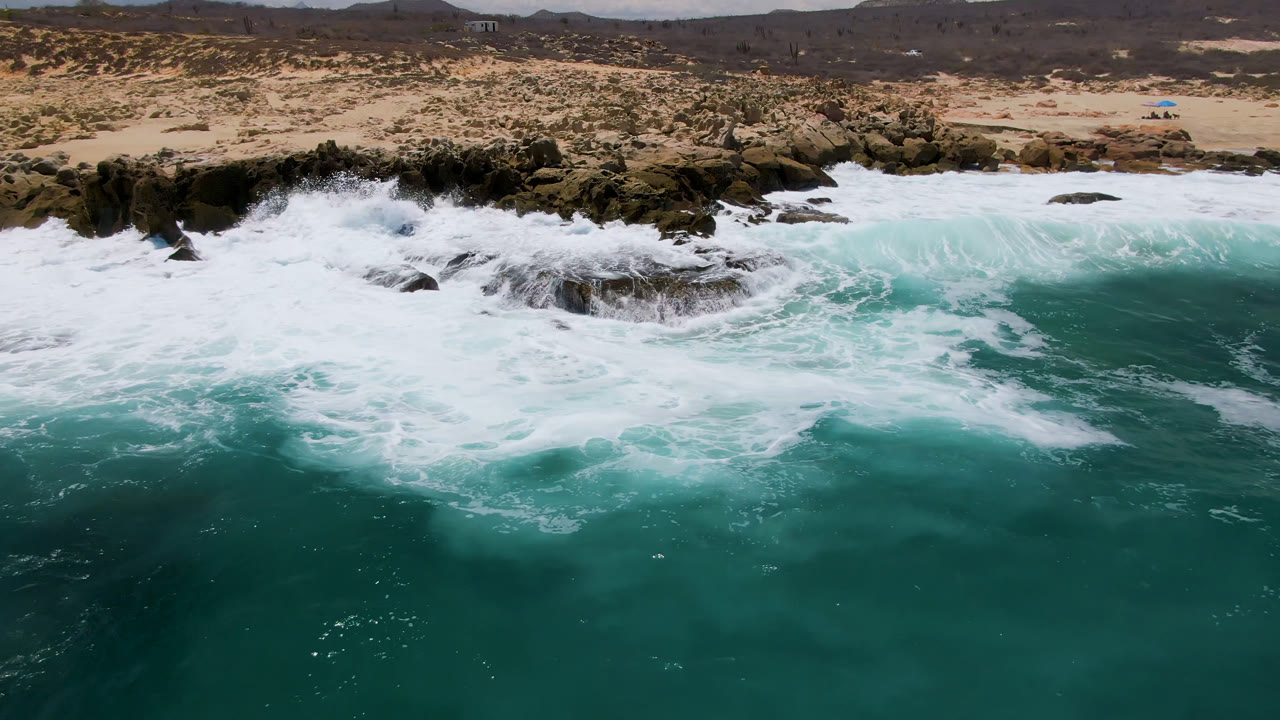 Aerial view at Laguna Beach, Baja California Sur, with turquoise water and white surf spreading over dark rocks below a sandy desert shoreline, with no visible people.