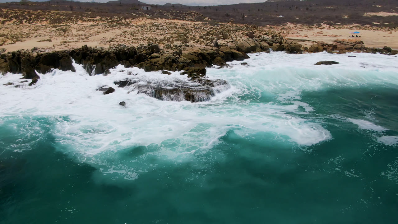 Foamy waves cover the nearshore rocks at Laguna Beach, Baja California Sur, while a tan beach and sparse scrubland stretch behind them, with no visible people.