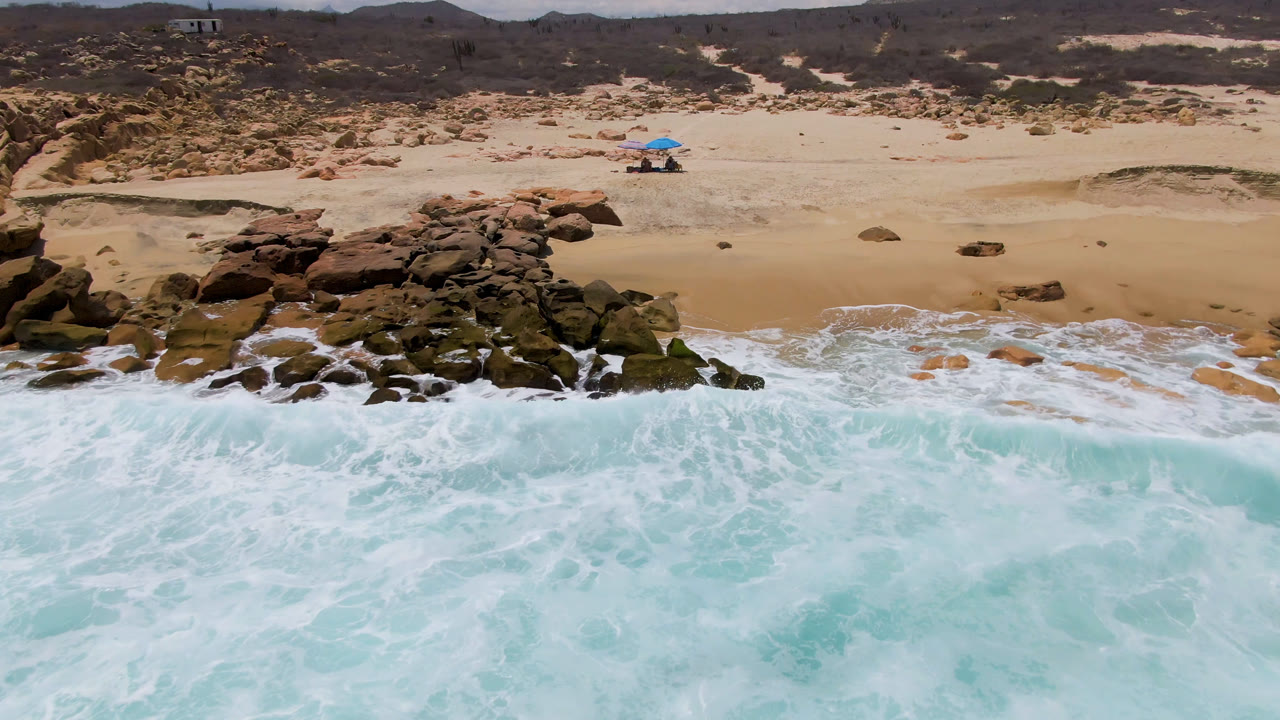 Aerial view of East Cape BCS beach in Mexico with foamy surf in the foreground, reddish rocks at left, and beach umbrellas set back on the sand.