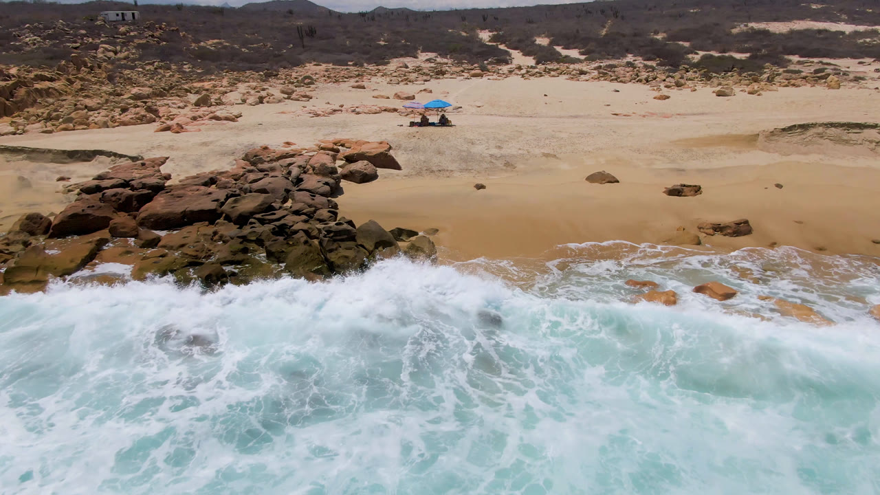 Drone-height view of East Cape BCS shoreline with whitewater spreading across turquoise water, a rock cluster near center, and umbrellas on the dry sand beyond.