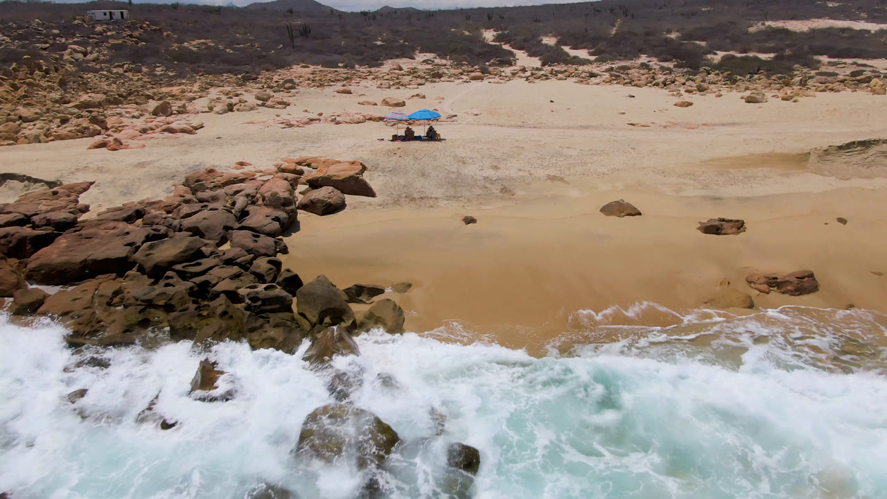 White surf crashes over rocks below beach umbrellas and dunes.