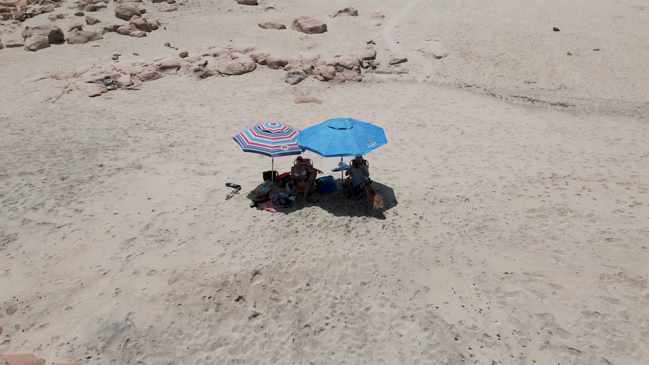 A high angle view in San Jose Del Cabo, Mexico shows the umbrella group centered low in the frame, with open sand above and rocky beach patches near the top edge.