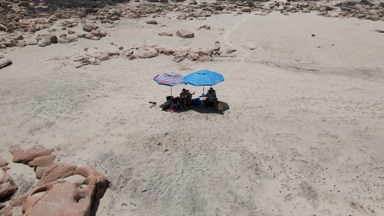 Aerial view of two umbrellas and seated beachgoers on pale sand near rocky shoreline.