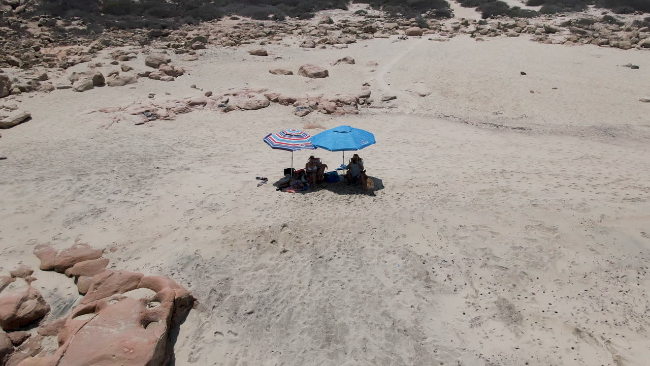 Aerial frame at 0:01 over Laguna Beach near San Jose Del Cabo, Mexico, showing the umbrella group slightly left of center, broad pale sand, rounded reddish rocks below, and sparse scrubland at the top.