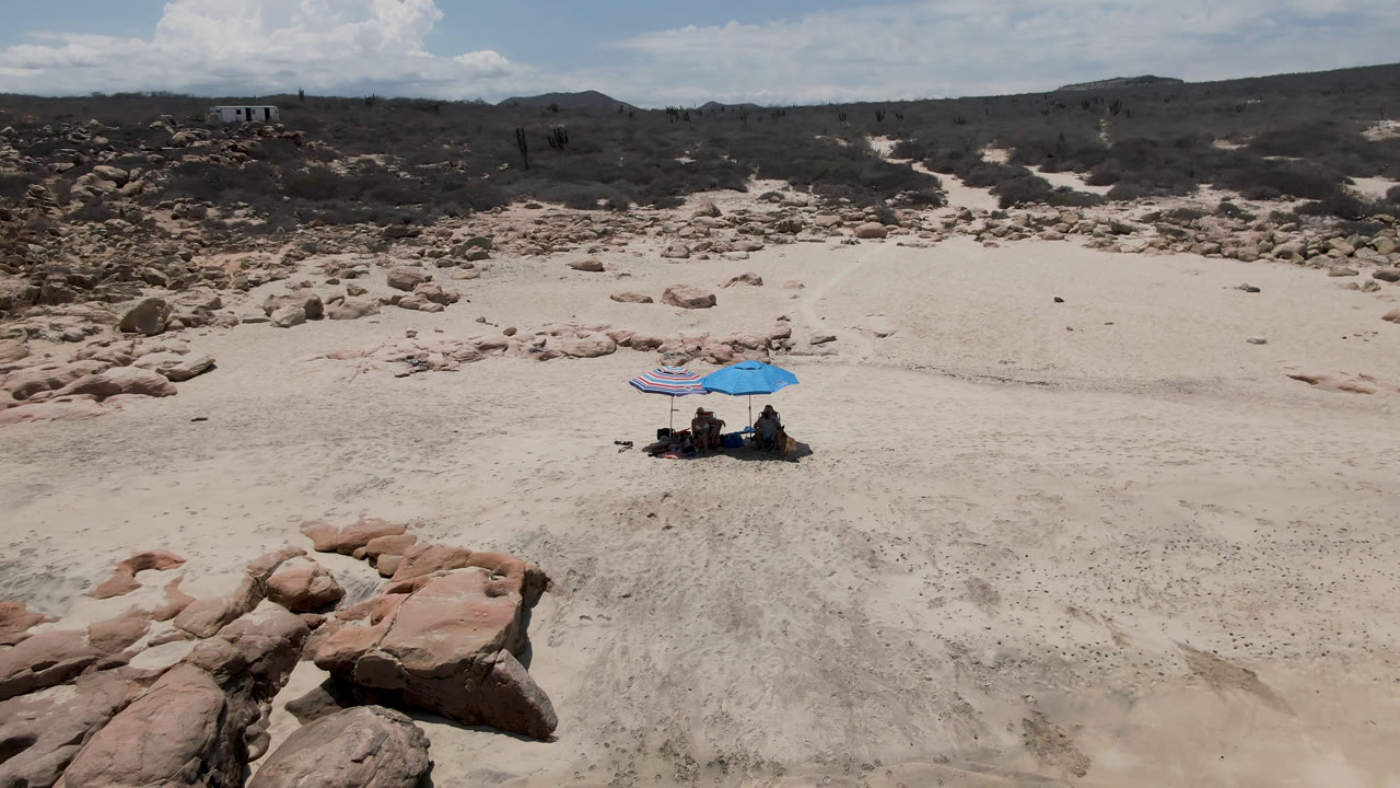 Elevated beach frame in San Jose Del Cabo, Mexico, with the umbrella group centered lower in the sand, reddish rocks closer to camera, and a dark scrub-covered ridge across the background.