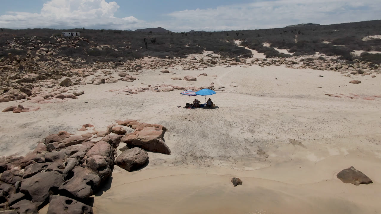 Wide aerial view of a sandy beach with rocks, dunes, and a blue umbrella.