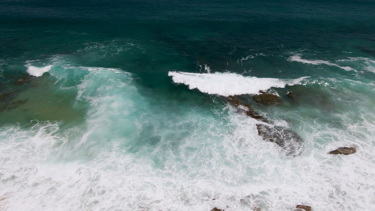 Aerial view of Laguna Beach, Baja California Sur, with white surf breaking over dark rocks and pale sand below, no visible people.