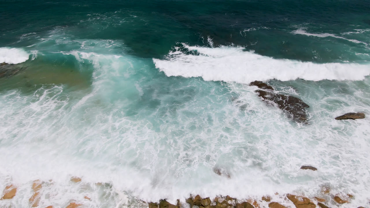 Overhead view at Laguna Beach, Baja California Sur, with whitewater wrapping around exposed rocks beneath darker green ocean, no visible people.