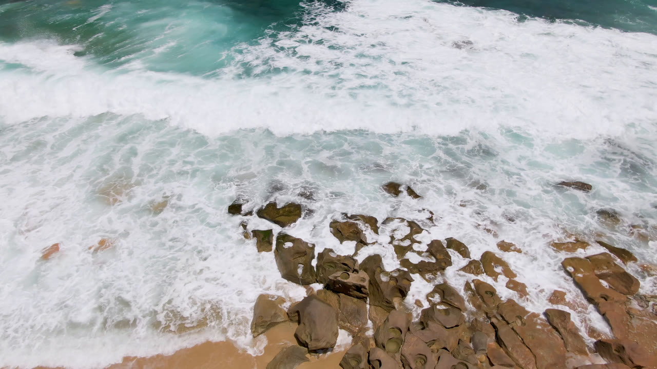 Overhead view of white surf washing over tan shoreline rocks and sand at Laguna Beach, Baja California Sur, with blue-green ocean above and no visible people.
