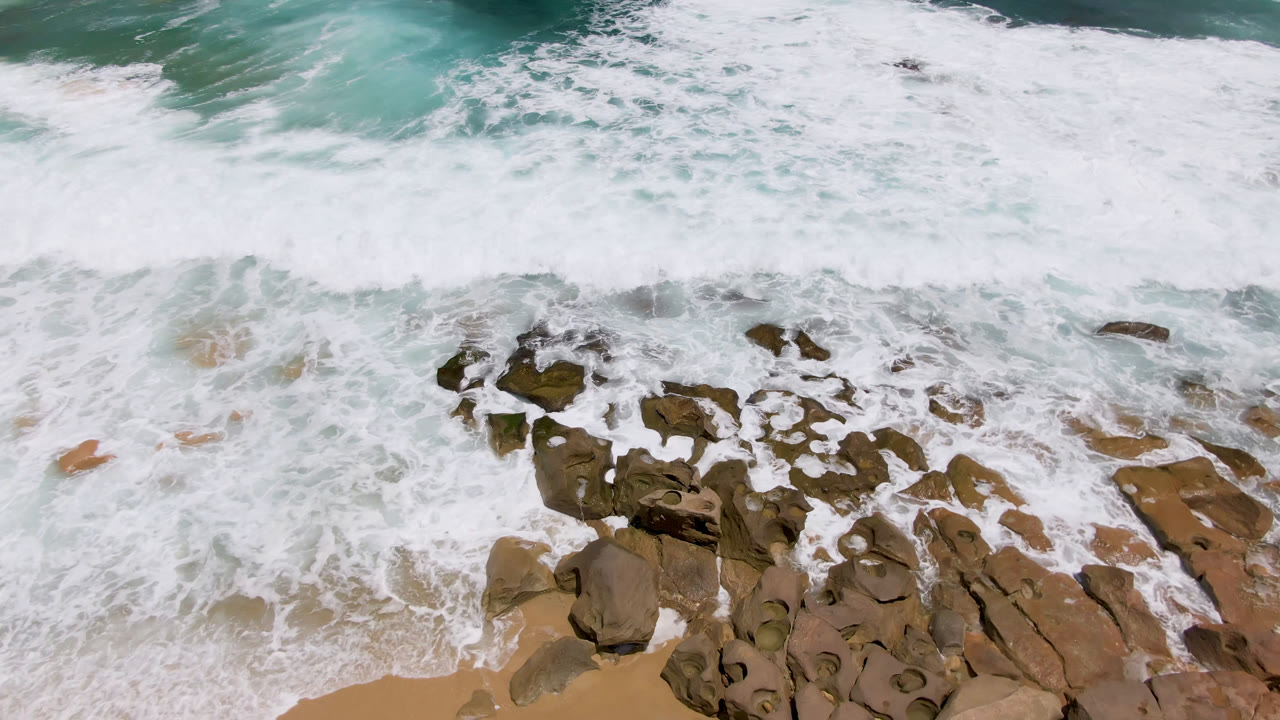 White foam spreads farther across clustered shoreline rocks at Laguna Beach, Baja California Sur, while deeper teal water fills the top of the frame and no visible people appear.