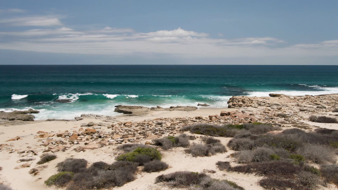 Laguna Beach coastline with more of the rock-strewn sand centered in frame, white water moving across shallow rocks, and a clear ocean horizon under scattered clouds.