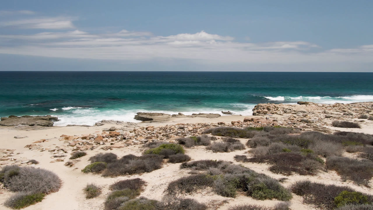 Wide beach and rocky shoreline with waves under a blue sky.