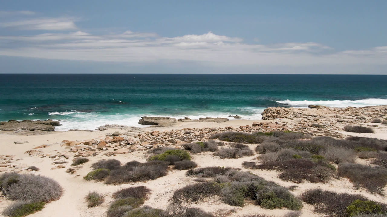 Laguna Beach shoreline with scrub patches across the sand, tan rocks near the waterline, white surf breaking offshore, and no visible people.