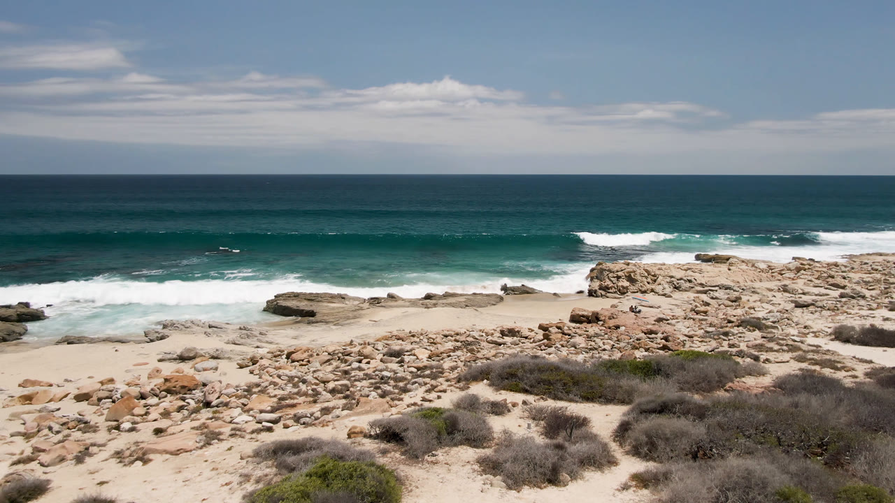 Rocky beach with surf, scrub, ocean horizon, and a small distant figure.