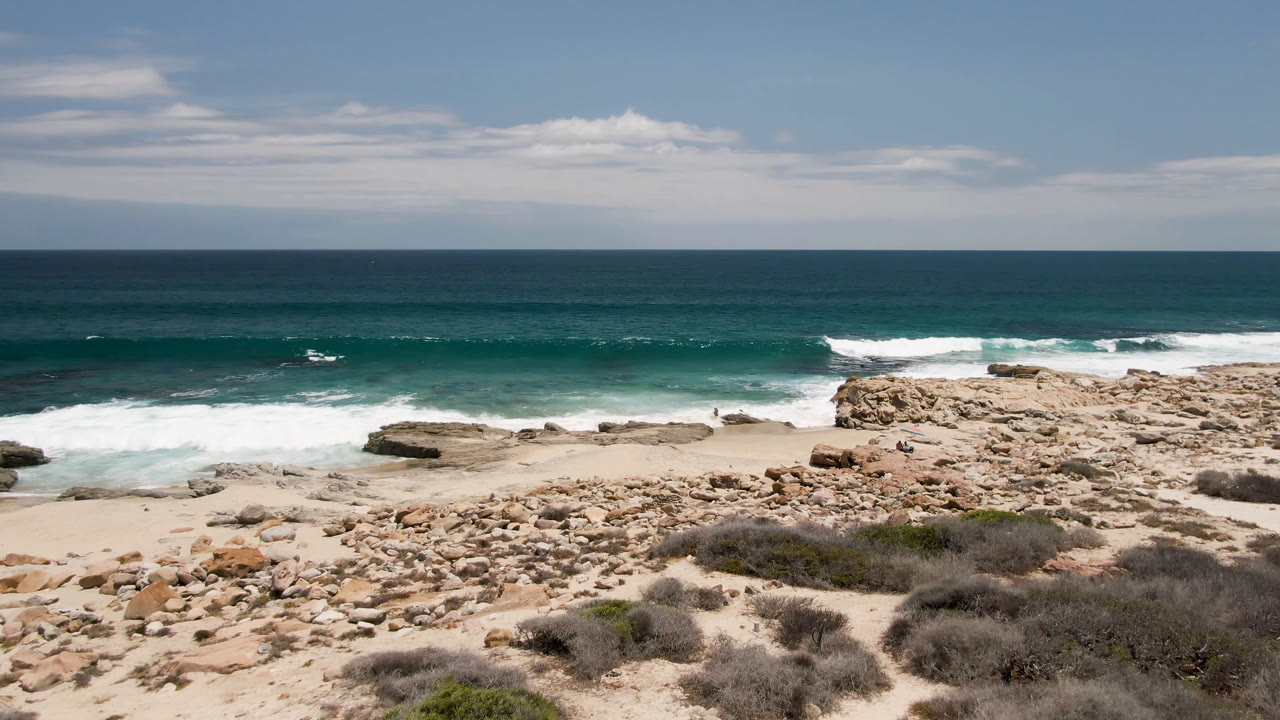 Laguna Beach shoreline in Baja California Sur with pale sand, jagged tan rocks, scrub in the foreground, white surf moving across the beach, and a small distant person on the rocks.