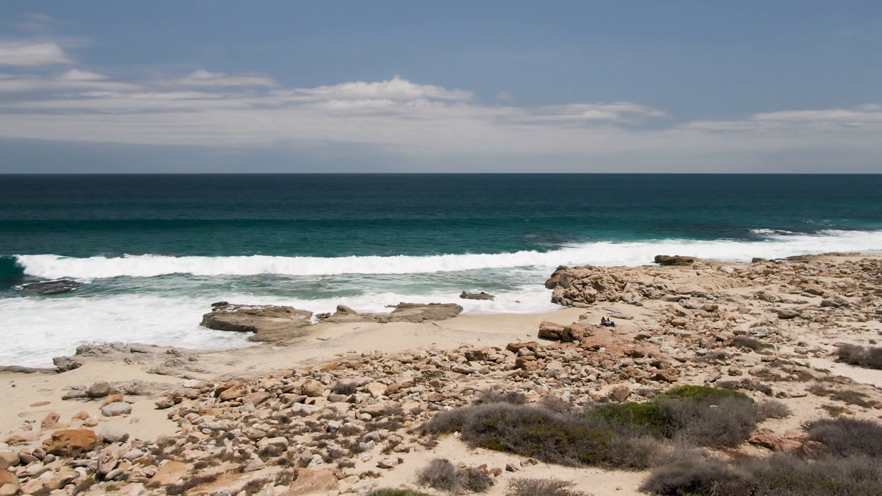 At Laguna Beach in Baja California Sur, Mexico, a rocky sandy shoreline fills the foreground as white waves break below a blue-green ocean horizon, with no visible people.