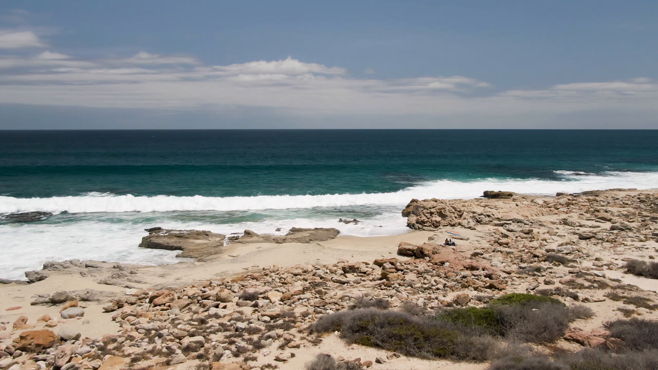 At Laguna Beach in Baja California Sur, Mexico, white surf pushes closer to the sandy cove beside a flat rock shelf, with open ocean and no visible people beyond it.