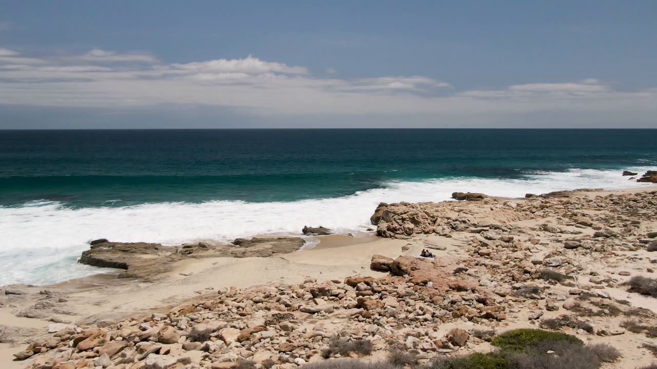 Opening frame of Laguna Beach in Baja California Sur, Mexico, showing tan rocks above a small sandy beach, white surf at the shore, blue-green ocean, and no visible people.