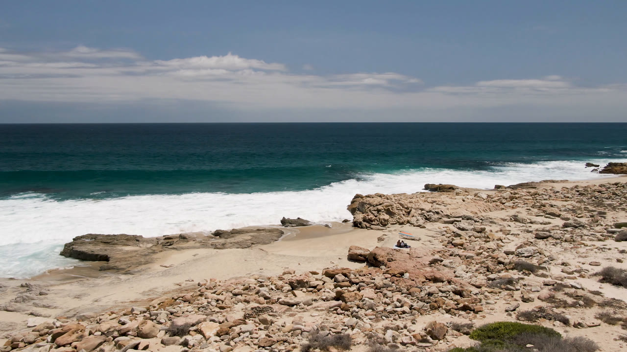 Laguna Beach shoreline in Baja California Sur, Mexico, with more of the central sandy pocket visible between scattered rocks, white foam, open ocean, and no visible people.