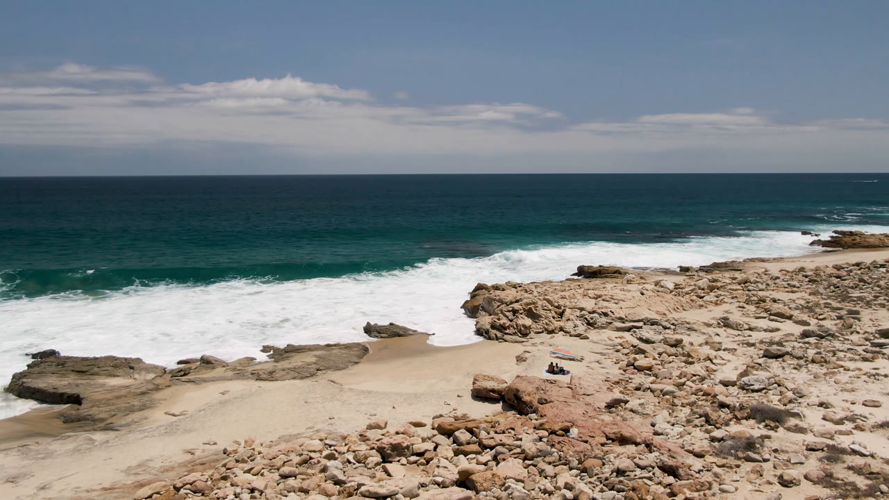 Wide elevated view of Laguna Beach in Baja California Sur, Mexico, with tan rocks in the foreground, white surf crossing the shore, open teal ocean, a cloud band, and tiny distant people near the sand.
